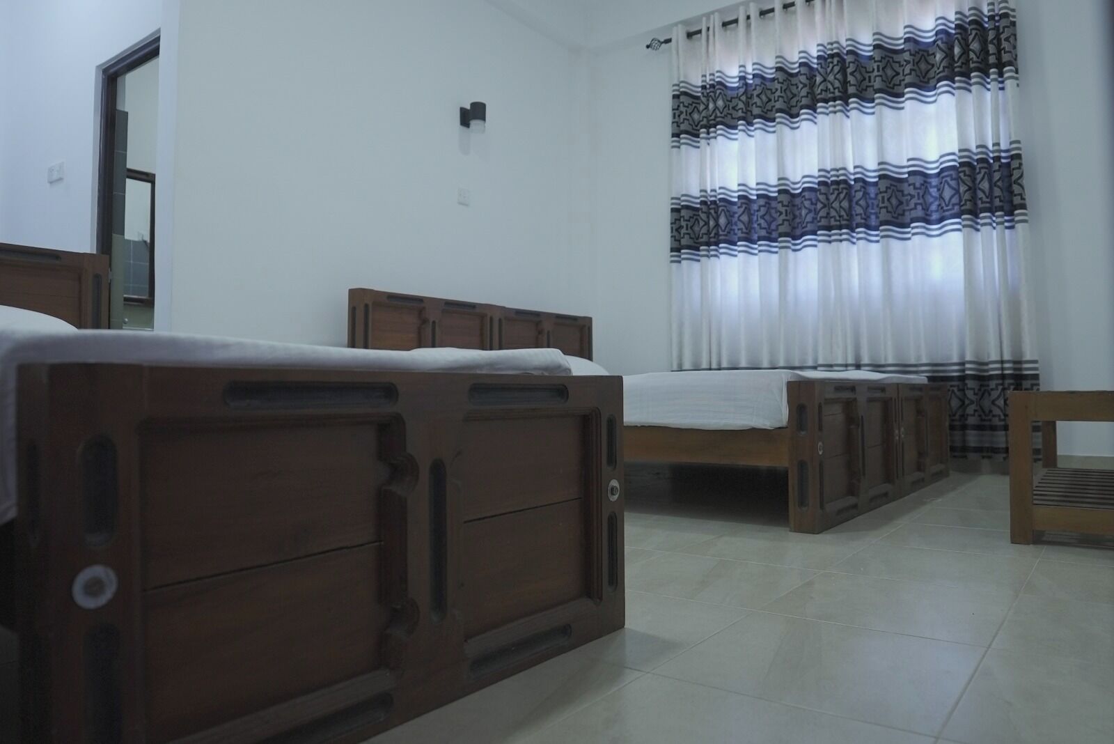 A bedroom with two wooden beds, a small wooden table, a wardrobe, and a window with patterned curtains.