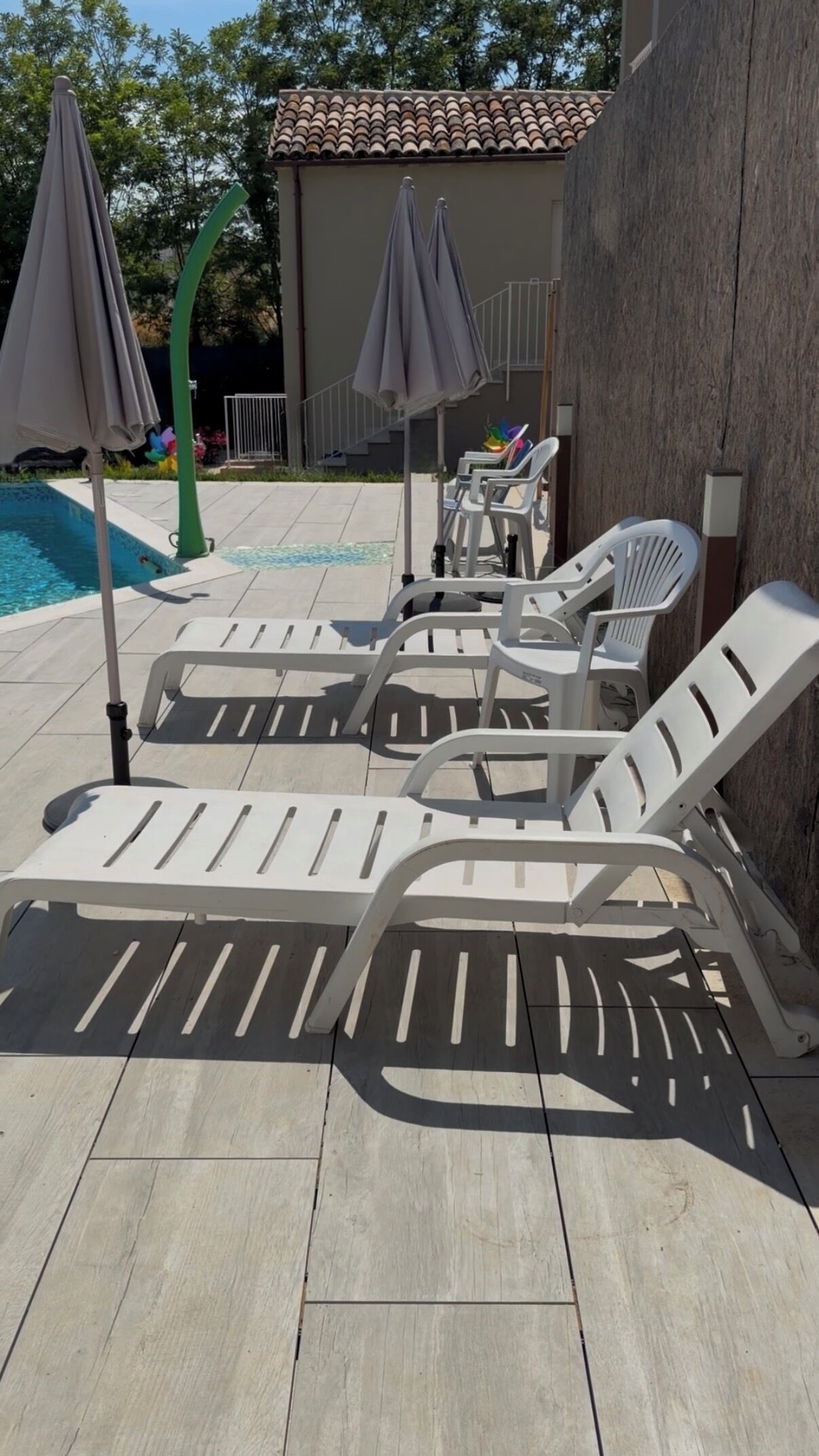 Ein Poolbereich mit Liegestühlen und Sonnenschirmen, ein gefliester Boden und im Hintergrund ein Gebäude mit einem Ziegeldach.
