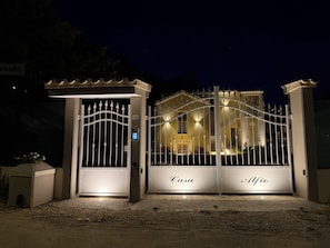 Een hekpoort met borden 'Casa' en 'Alfio', een beveiligingscodeklavier en een goed verlichte gevel in de nacht.