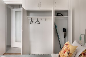 Ein weißer Kleiderschrank mit Kleiderstange, ein Bett mit gemustertem Kissen und ein Nachttisch mit Lampe.