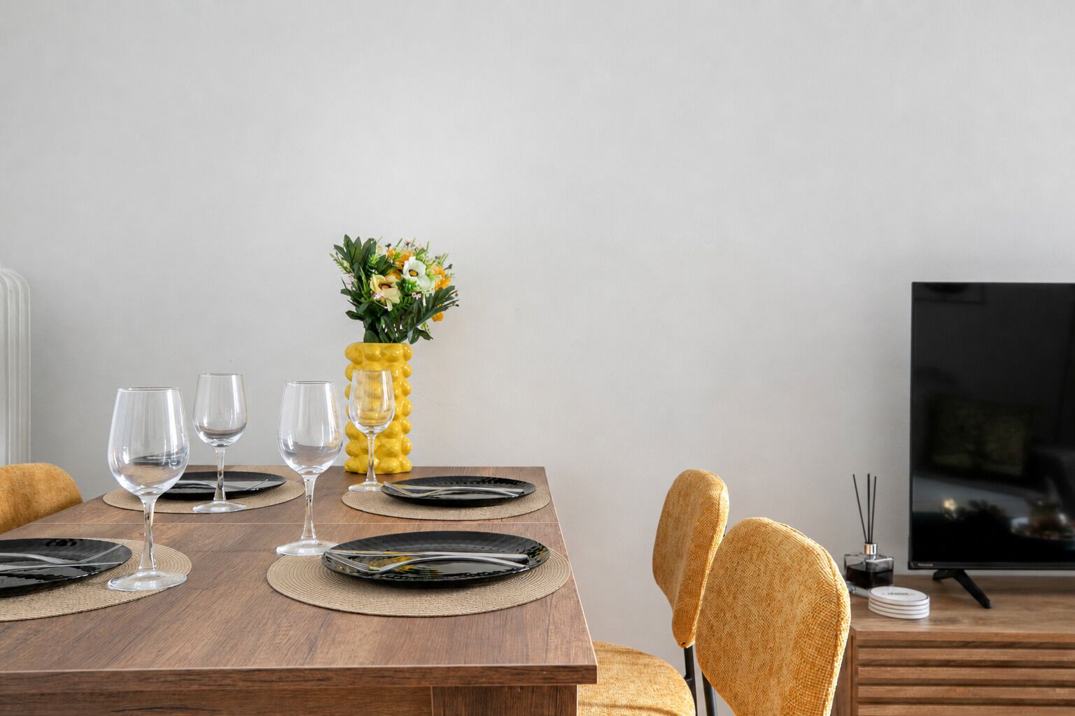 Una mesa de comedor con platos negros, copas de vino transparentes y un jarrón amarillo con flores.
