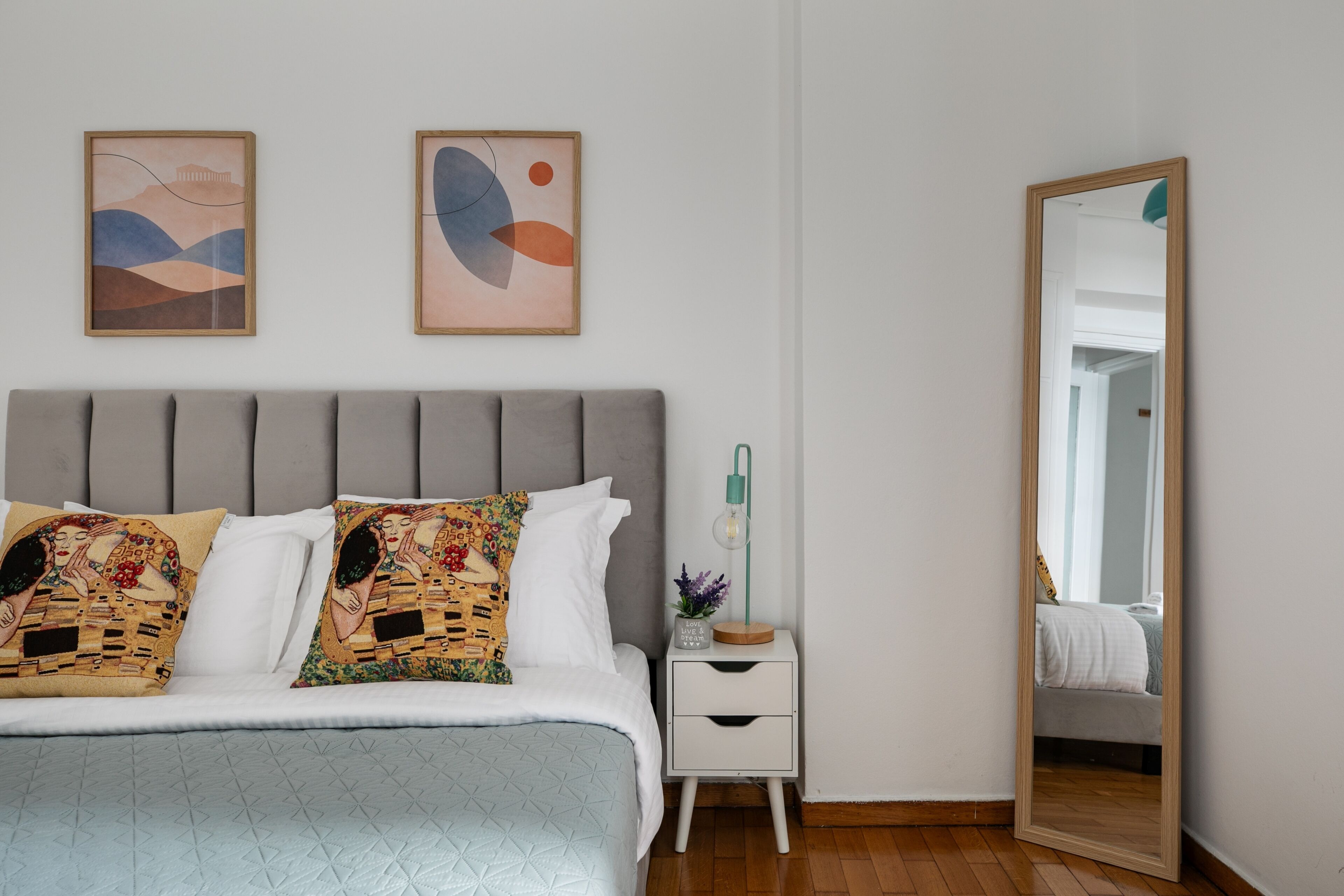 Une chambre à coucher comprenant un lit, une table de chevet, un miroir et deux œuvres d’art abstraites accrochées au mur.