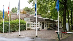 A building with a covered entrance, flags, and a paved area with a bicycle. - Gezellig huisje aan de bosrand in Rijen (Rijen)