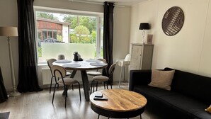 A modern living room with a round wooden coffee table, a black sofa, a dining table with chairs, and a view of greenery outside. - Gezellig huisje aan de bosrand in Rijen (Rijen)