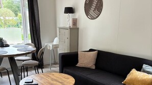 A modern living room with a round wooden coffee table, a black sofa with cushions, a dining area with a white table and chairs, and a decorative wall piece. - Gezellig huisje aan de bosrand in Rijen (Rijen)