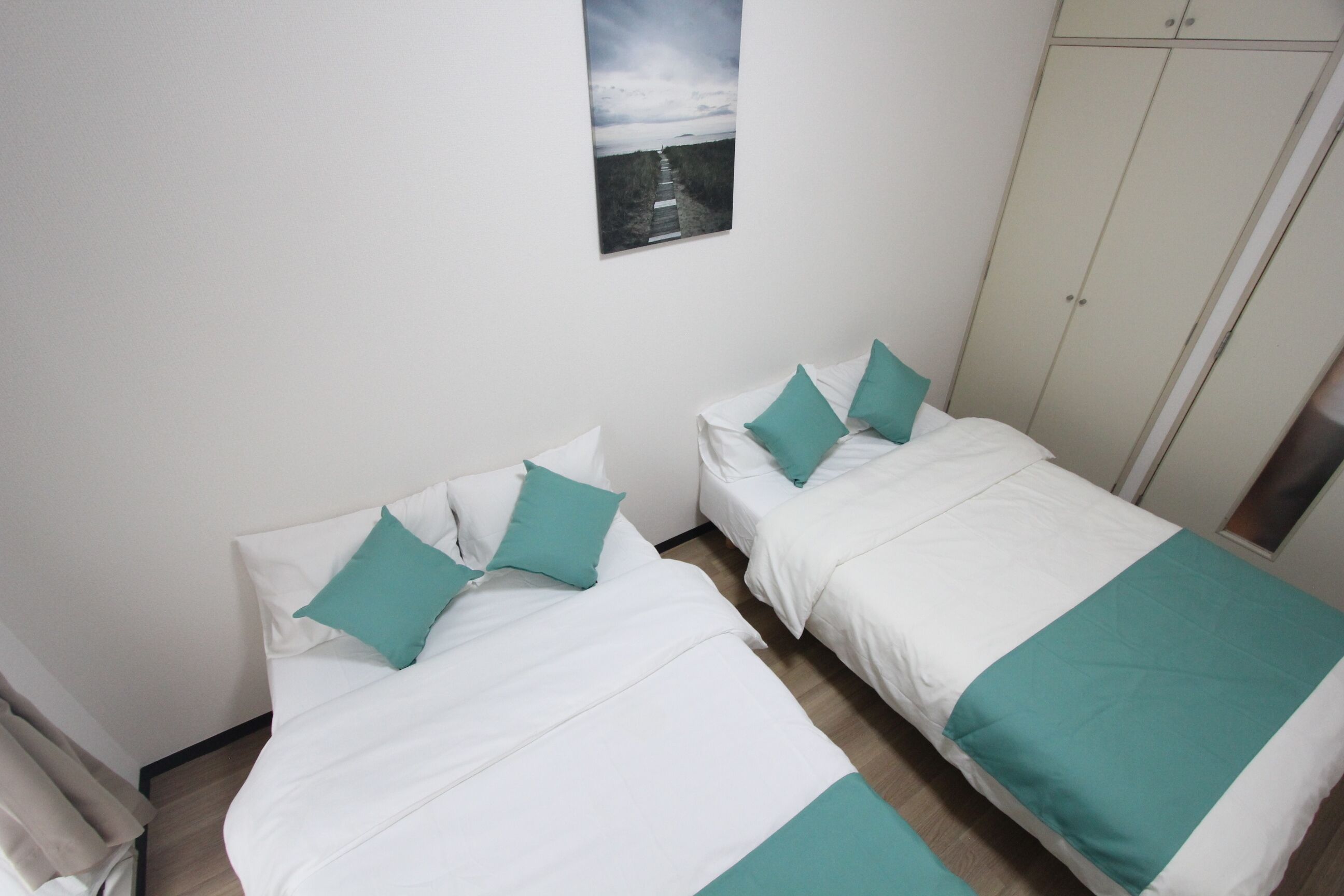 A room with two beds, each with white bedding and teal pillows, a white wardrobe, and a framed picture on the wall.