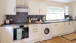 A modern kitchen with white cabinets, a built-in oven, a washing machine, and a window with blinds. - Laundry Cottage (Strathpeffer)