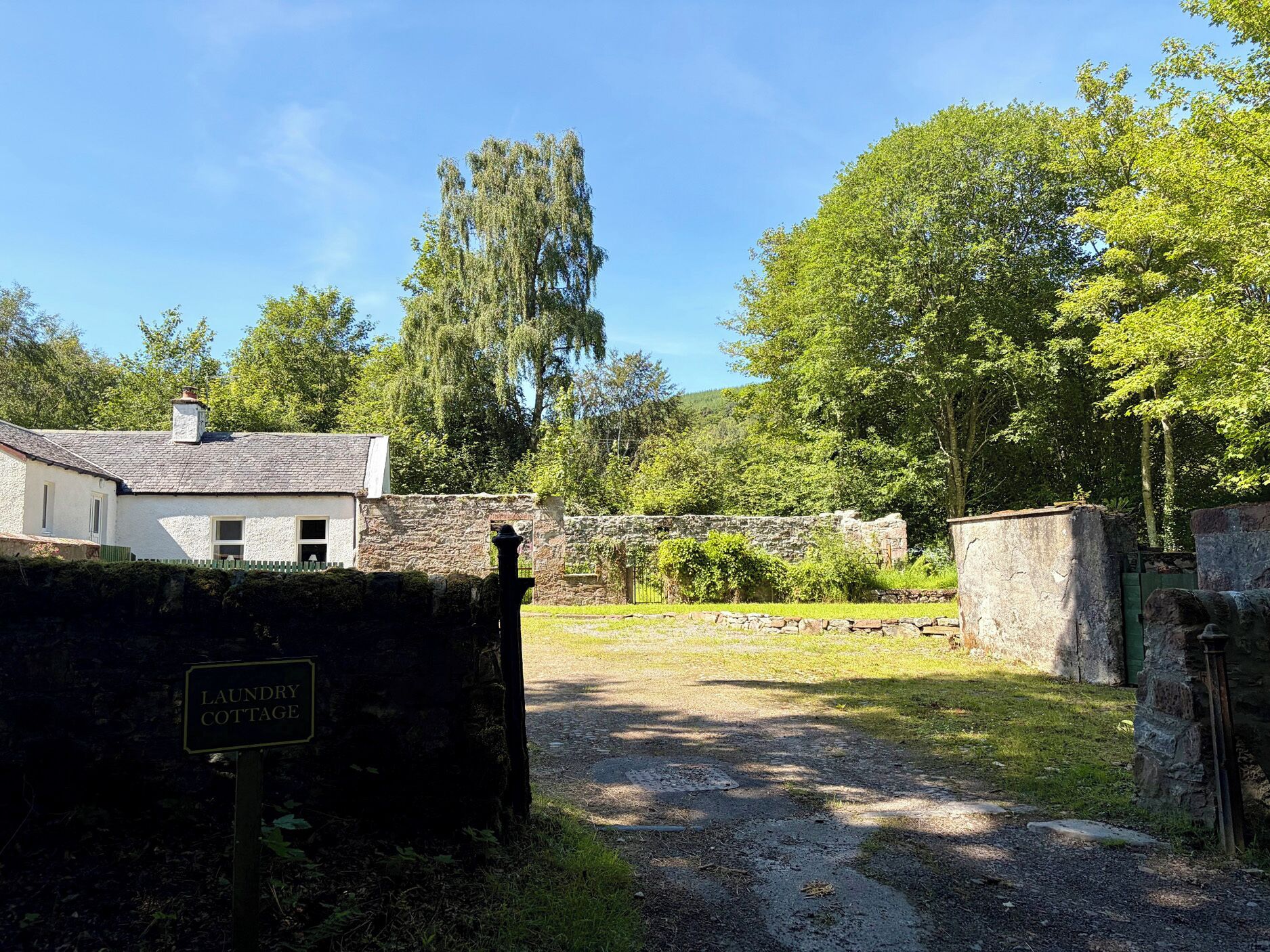 Een landelijk gelegen pand met een stenen muur, een bord waarop 'LAUNDRY COTTAGE' staat, en een grindpad dat naar een gazon leidt.