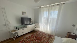 A bedroom with a bed, a TV on a white cabinet, a chair, a desk, and a window with curtains.