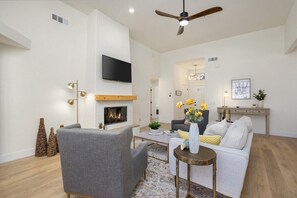 A modern living room with a fireplace, a ceiling fan, a flat-screen TV, and a dining area with a coffee table. - The Refuge - Desert Escape w/ Pool Spa & Game Room (Desert Hot Springs)