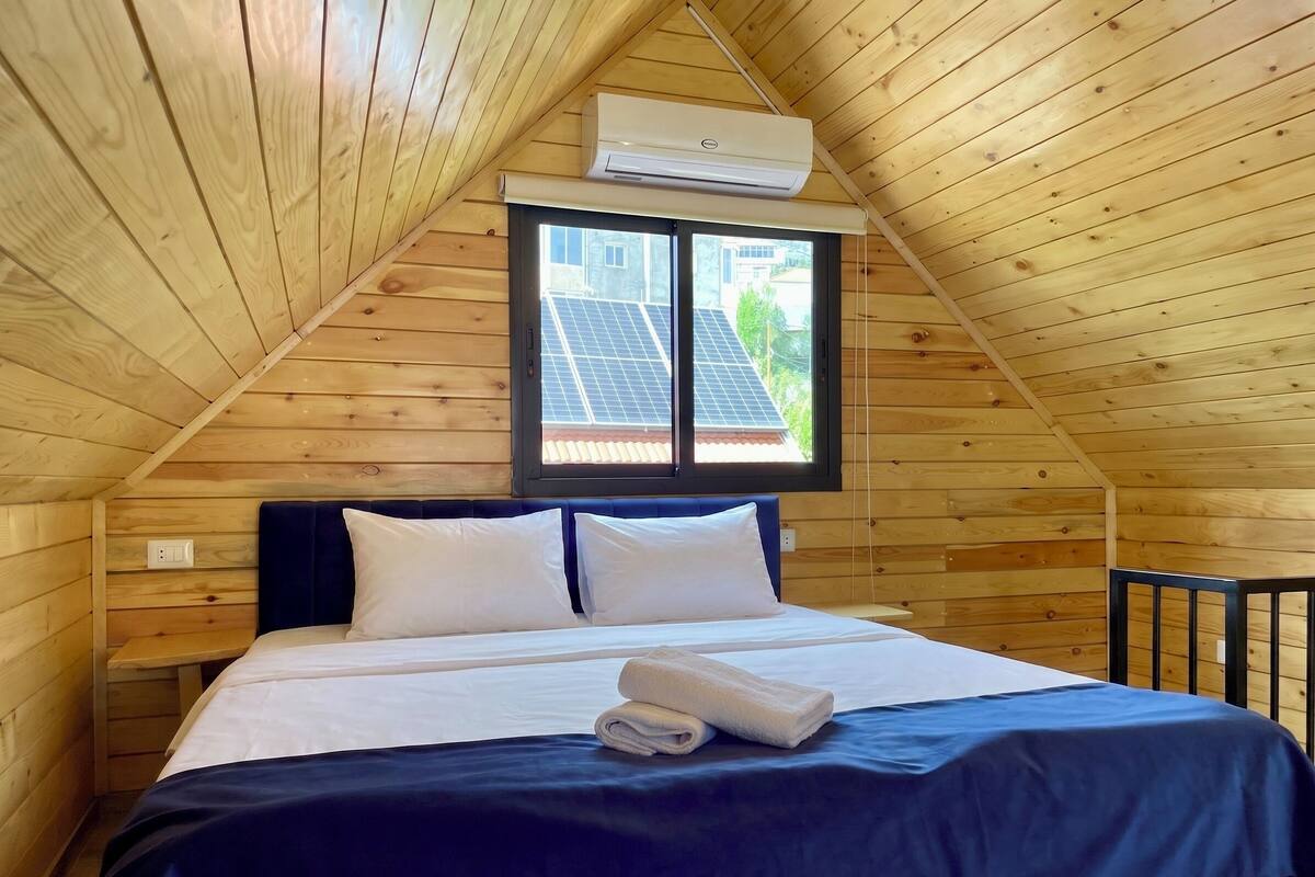 Una habitación de madera con una cama, un ventanal con paneles solares y un equipo de aire acondicionado.
