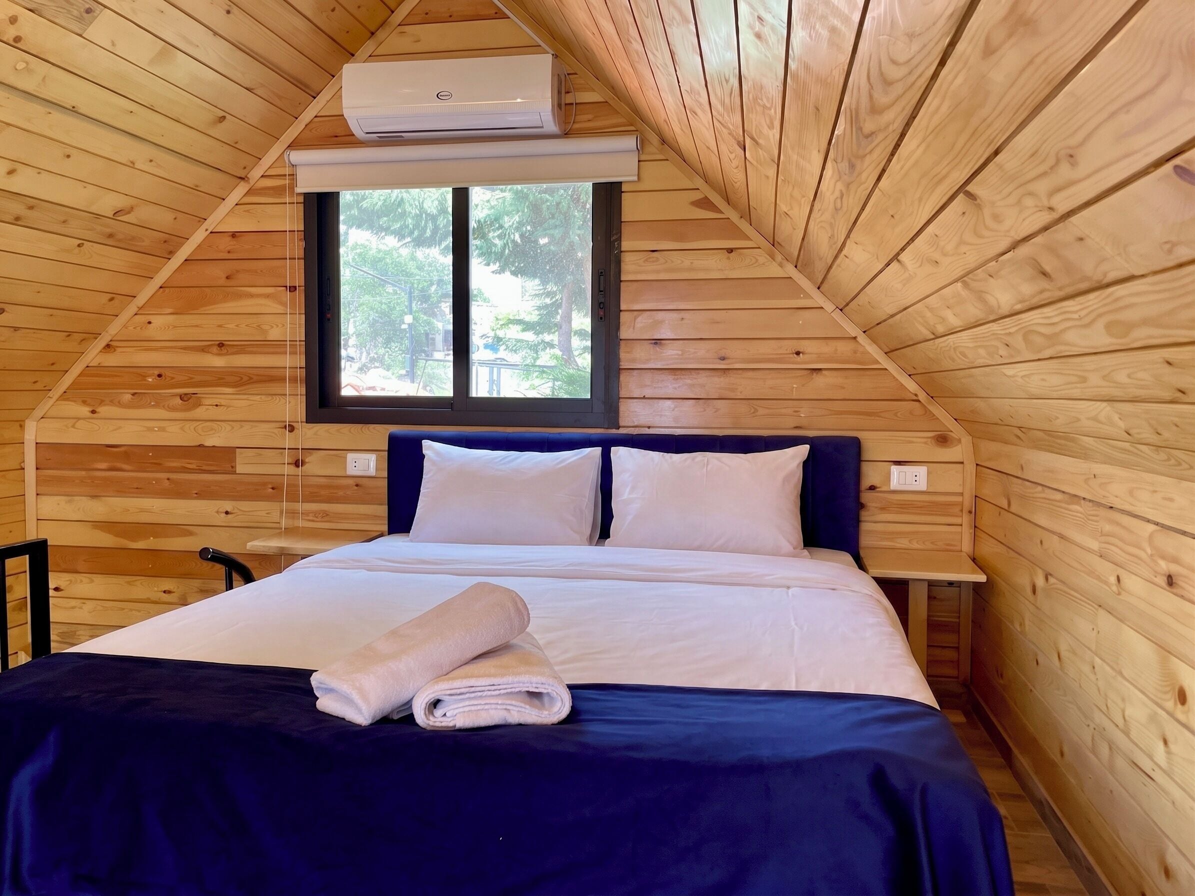 A bedroom with a wooden ceiling, a window, a bed with white bedding, and a rolled towel on the bed.