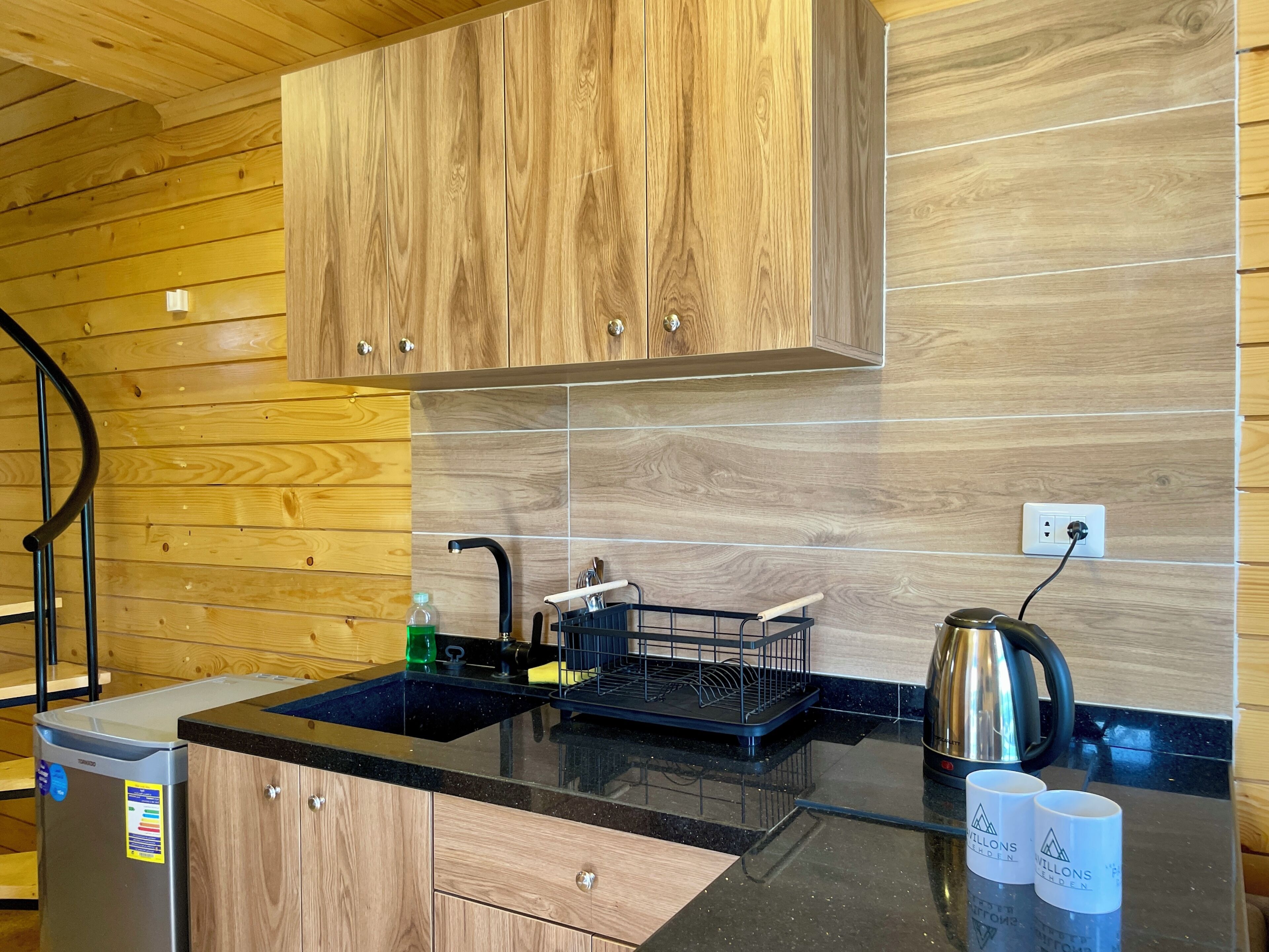 A modern kitchen with wooden cabinets, a black countertop, a stainless steel kettle, and a dish rack.
