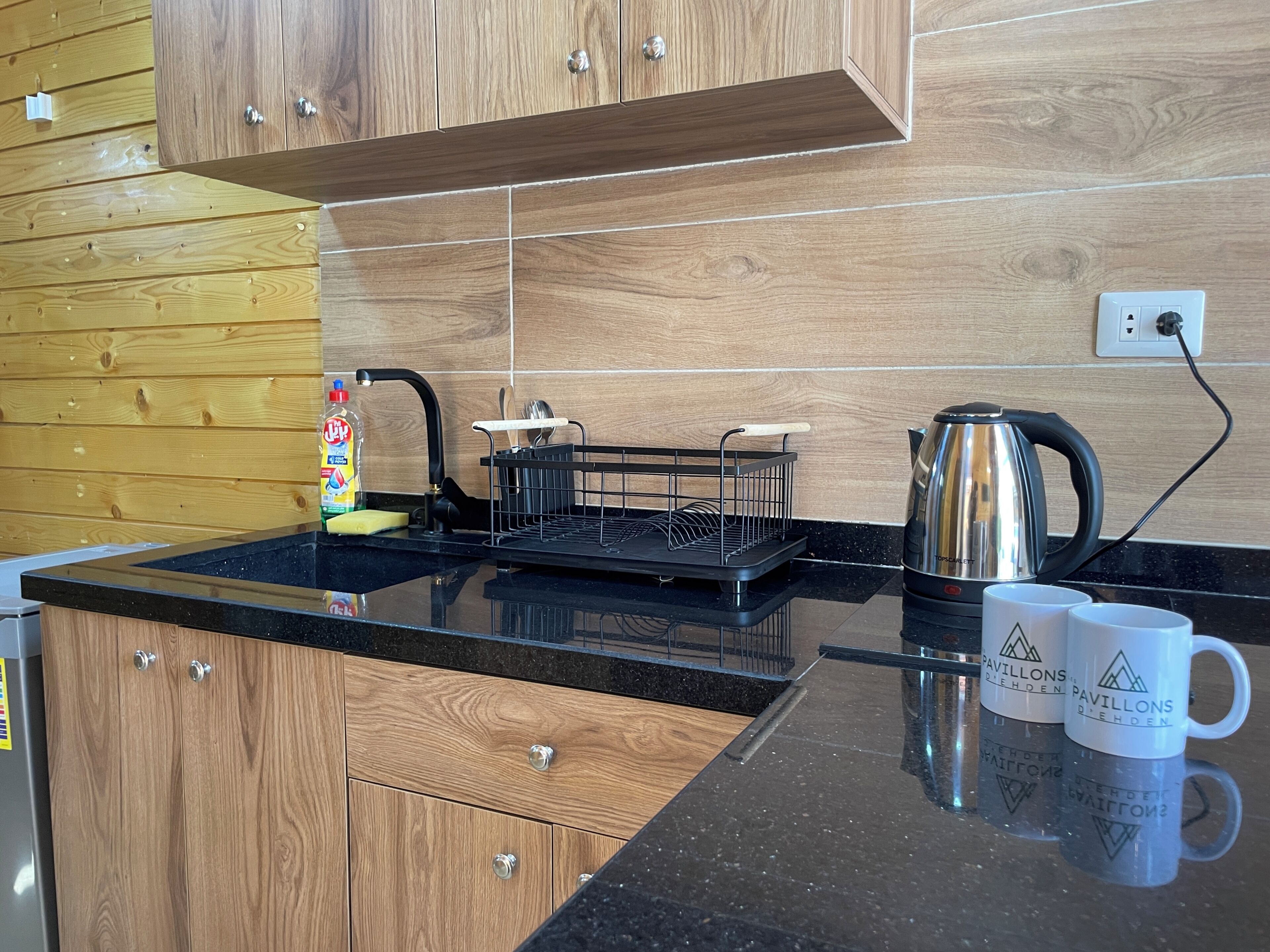 A modern kitchen with wooden cabinets, a black countertop, a kettle, and two mugs.