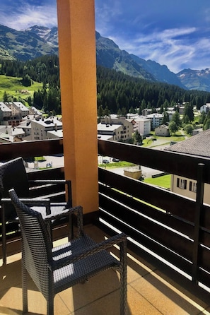 Un balcon avec des fauteuils en osier, d’où l’on peut admirer une ville avec ses bâtiments et des montagnes en arrière-plan.
