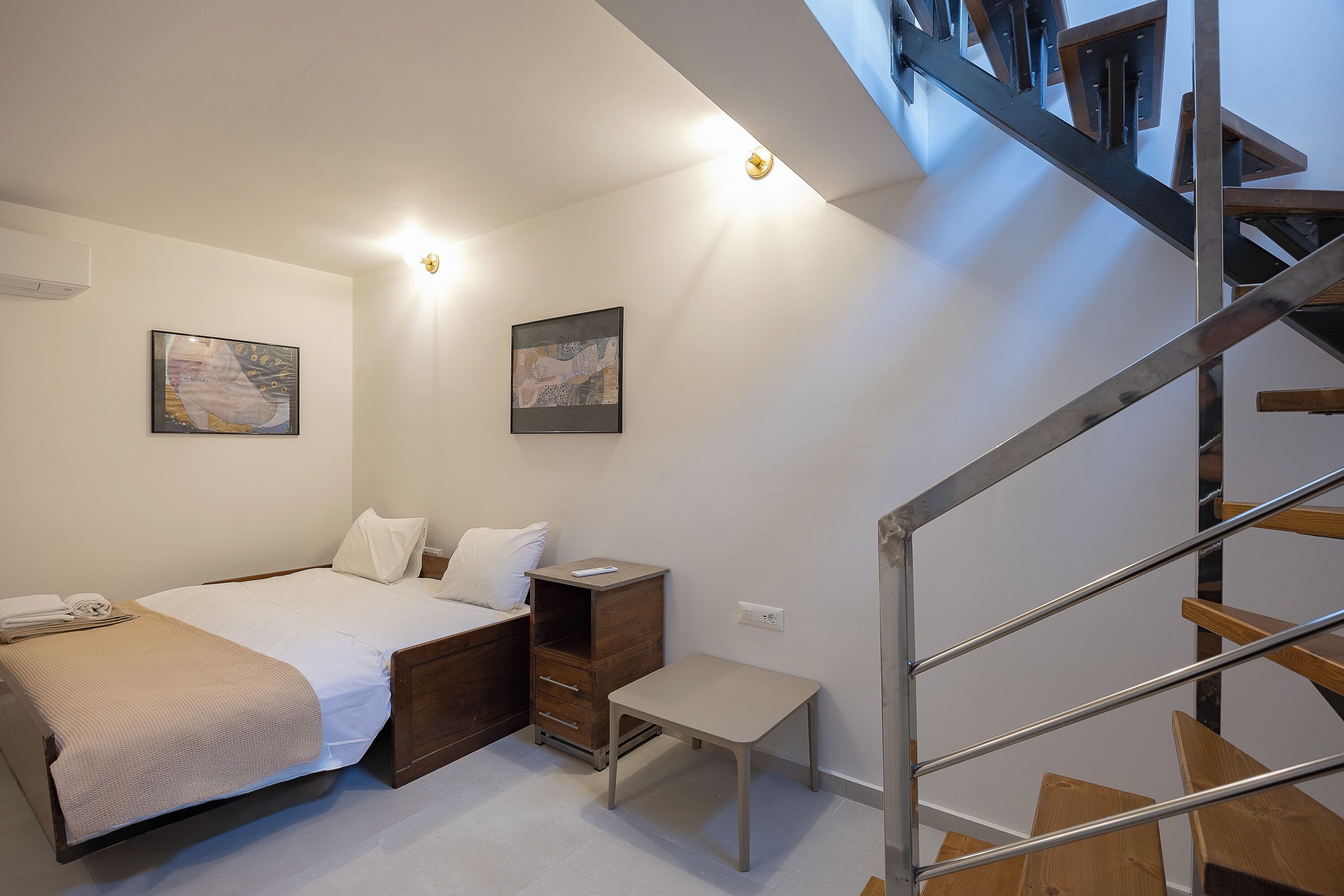 Une chambre à coucher avec un lit, une table de chevet et une petite chaise. Un escalier avec des rampes en métal mène à un niveau supérieur.