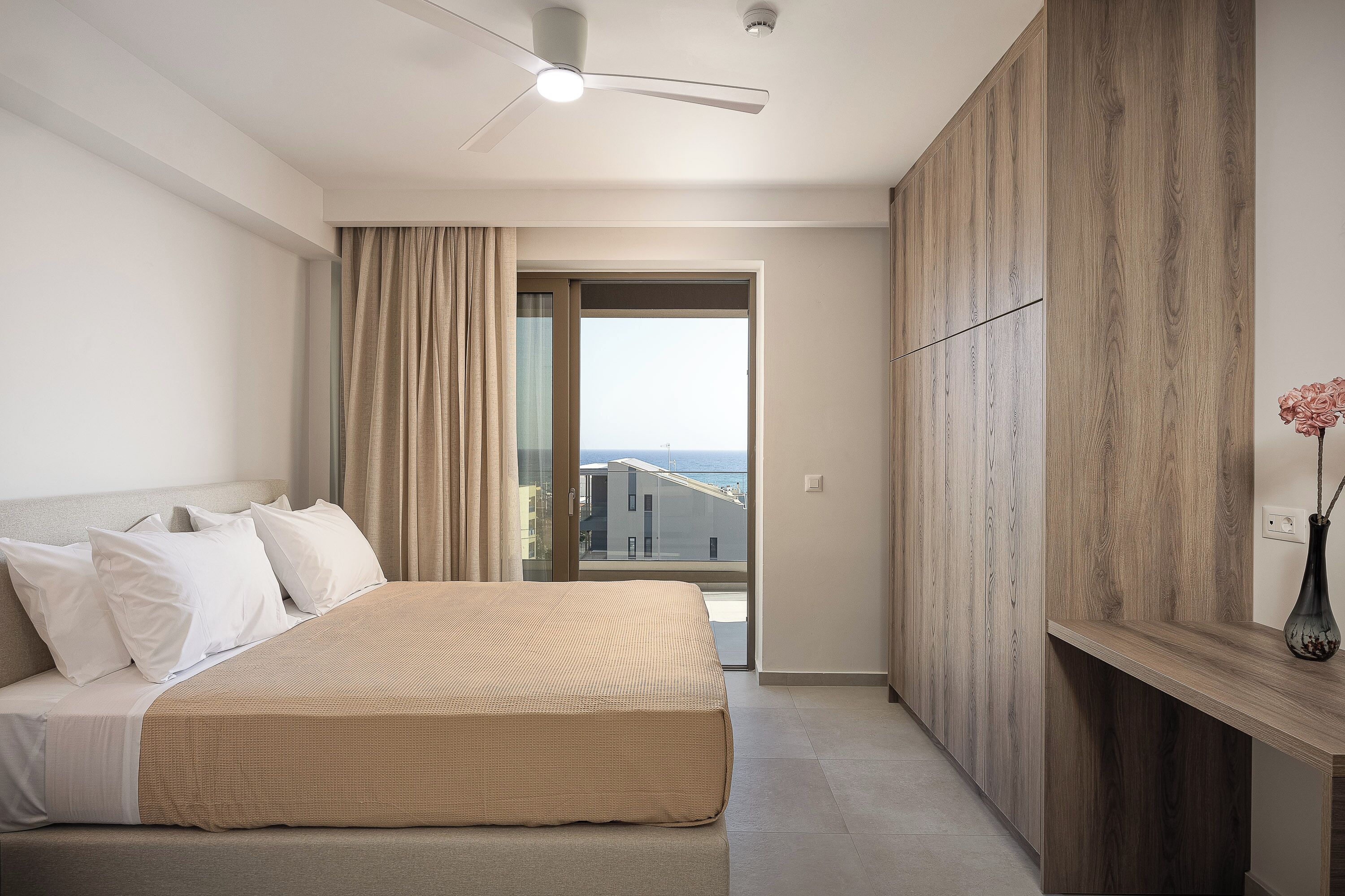 Habitación de hotel con cama, ventilador de techo, vista por una puerta de vidrio y un armario de madera.