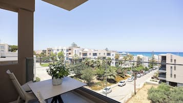 A balcony with a table and chairs, overlooking a residential street with cars and buildings, and a clear view of the sea.