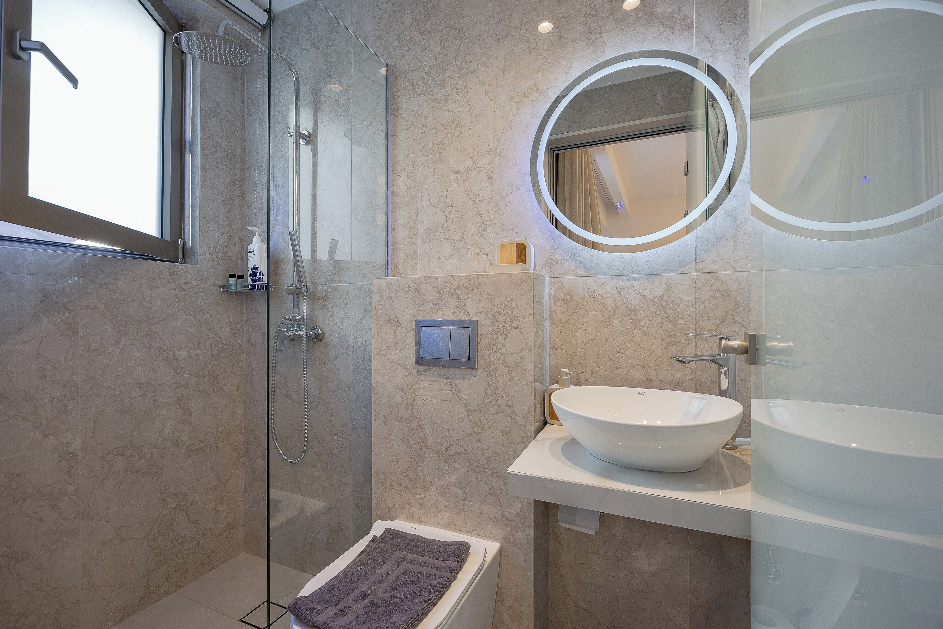 A modern bathroom with a glass-enclosed shower, a round mirror, and a white sink.