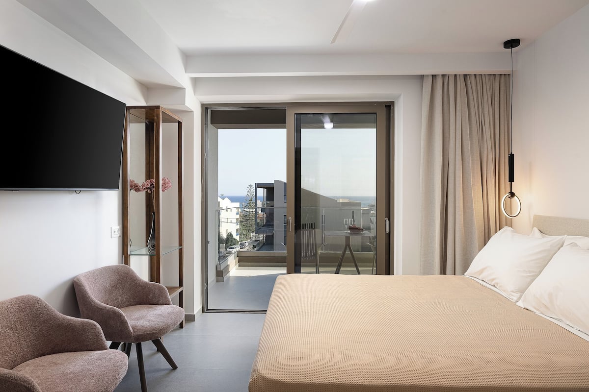 Ett modernt hotellrum med en säng, två fåtöljer, en TV och en balkong med utsikt över staden.