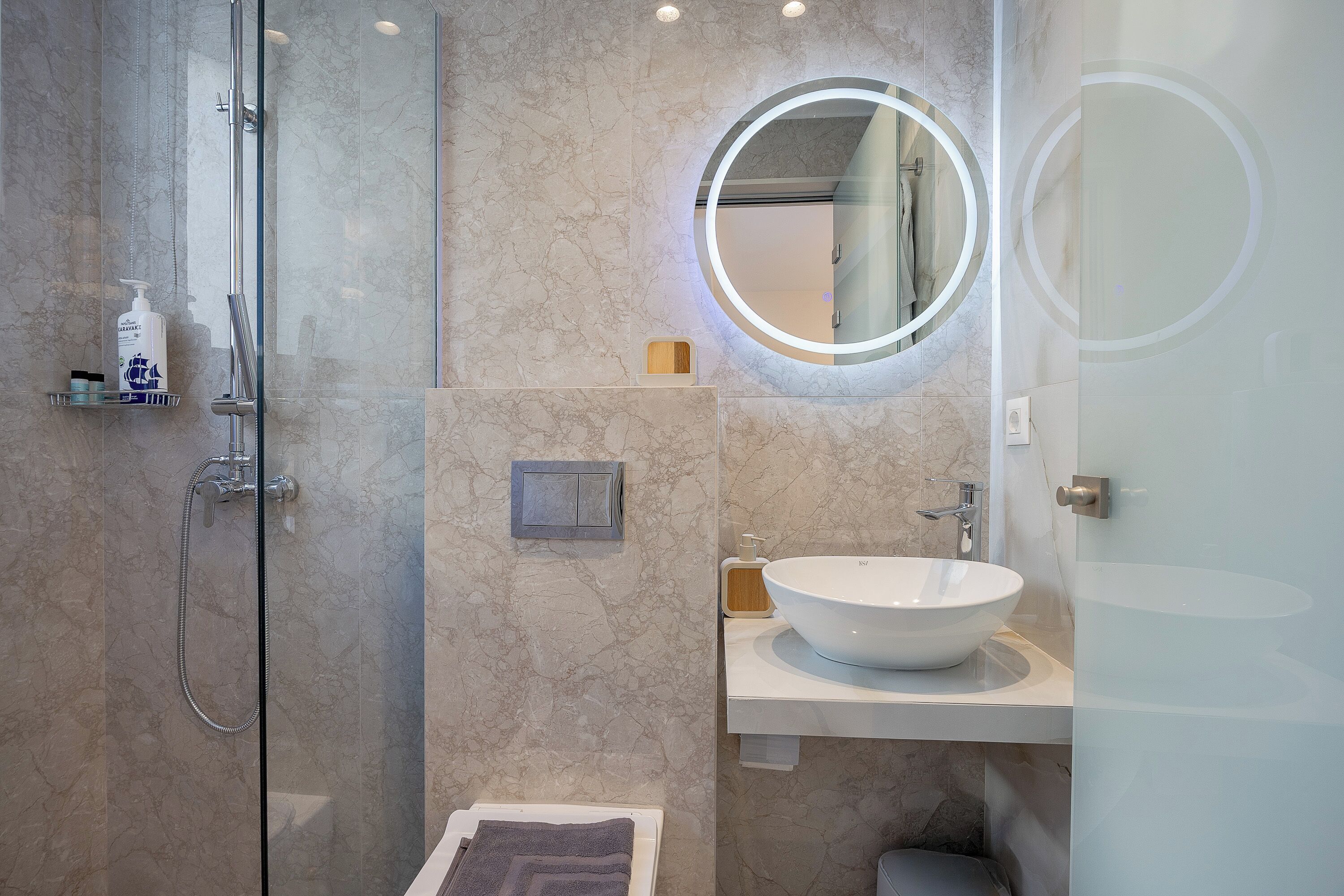 Un baño moderno con ducha envuelta en vidrio, un espejo redondo y un lavamanos blanco.