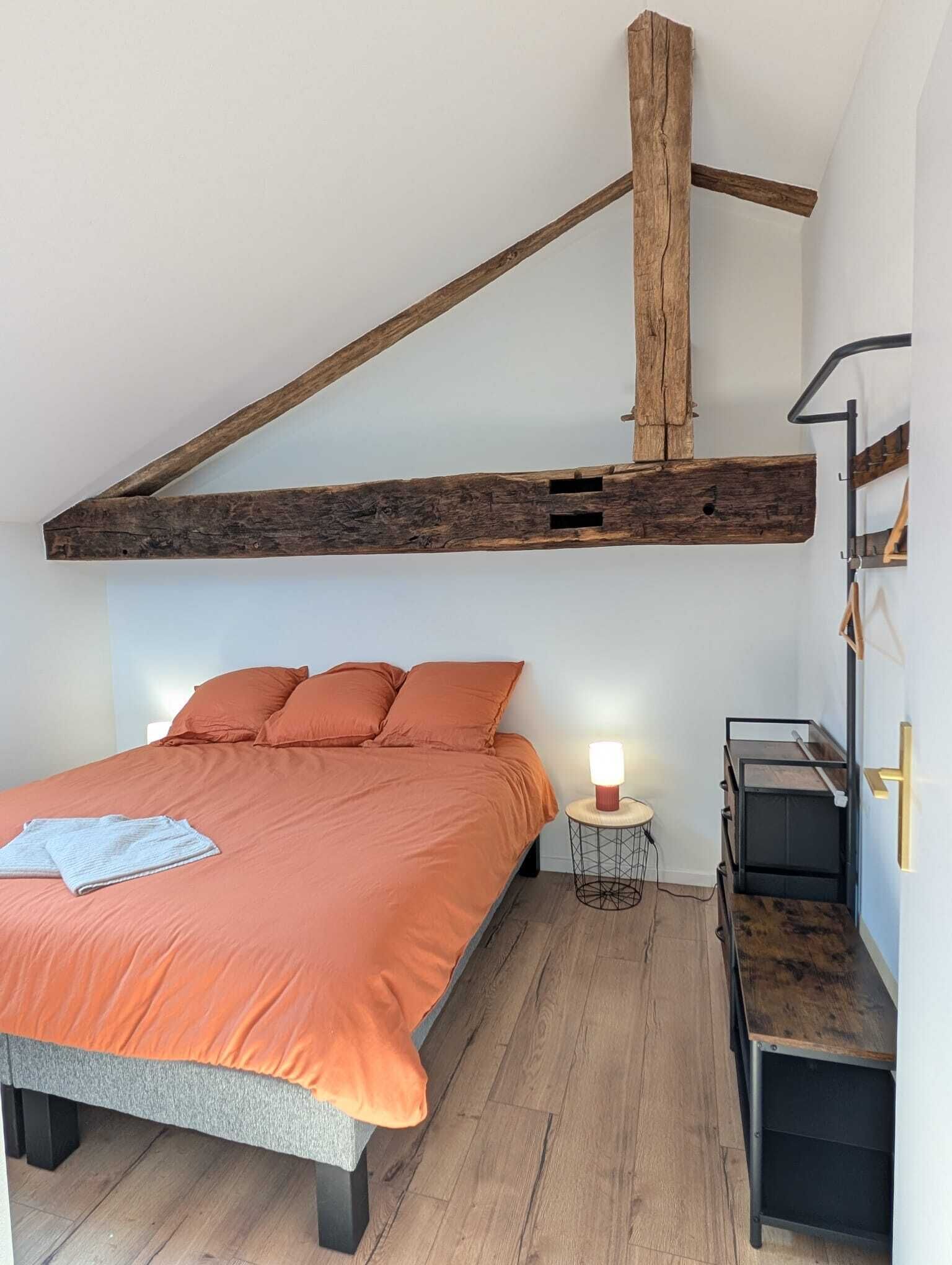 Un dormitorio con techo inclinado, una cama con ropa de cama naranja, una mesita de noche con una lámpara y un pequeño banco de madera.