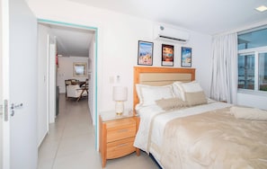 Habitació d'hotel amb llit, taula de nit, làmpada i aire condicionat fixat a la paret. Hi ha quadres emmarcats a la paret i una finestra amb cortines.