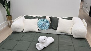 Ein Zimmer mit einem grünen Sofa, weißen und dunkelgrünen Kissen, einem blauen Zierkissen und einem aufgerollten weißen Handtuch auf dem Boden.
