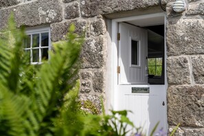 A stone building with a white door and window, surrounded by greenery. - Tinners Cottage, Pendeen – Sleeps 2 – Garden – Woodburner (Pendeen)