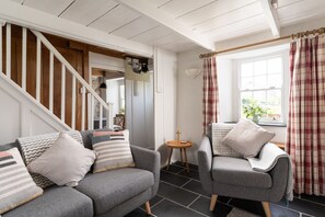 A living room with a grey sofa, a grey armchair, a small round table, a staircase, and a window with curtains. - Tinners Cottage, Pendeen – Sleeps 2 – Garden – Woodburner (Pendeen)