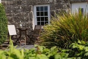 A stone building with a white door, a window, and a table with two chairs. - Tinners Cottage, Pendeen – Sleeps 2 – Garden – Woodburner (Pendeen)
