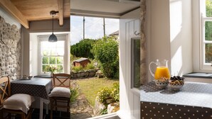 Ein Zimmer mit Stehwänden, Holzbalkendecke, einem Tisch mit Tischdecke mit Punkten und einem Fenster mit Gartenblick.