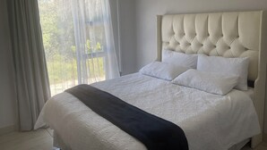 A bedroom with a large bed, a window with sheer curtains, and an air conditioning unit.
