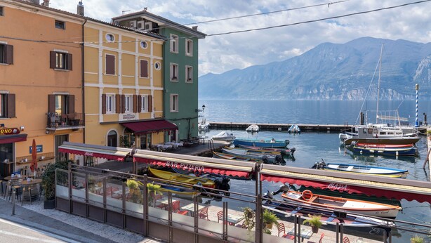 A waterfront view with boats docked, buildings with balconies, and a mountain backdrop. - Apartment with a lake view in the center of Castelletto di Brenzone (Castelletto)