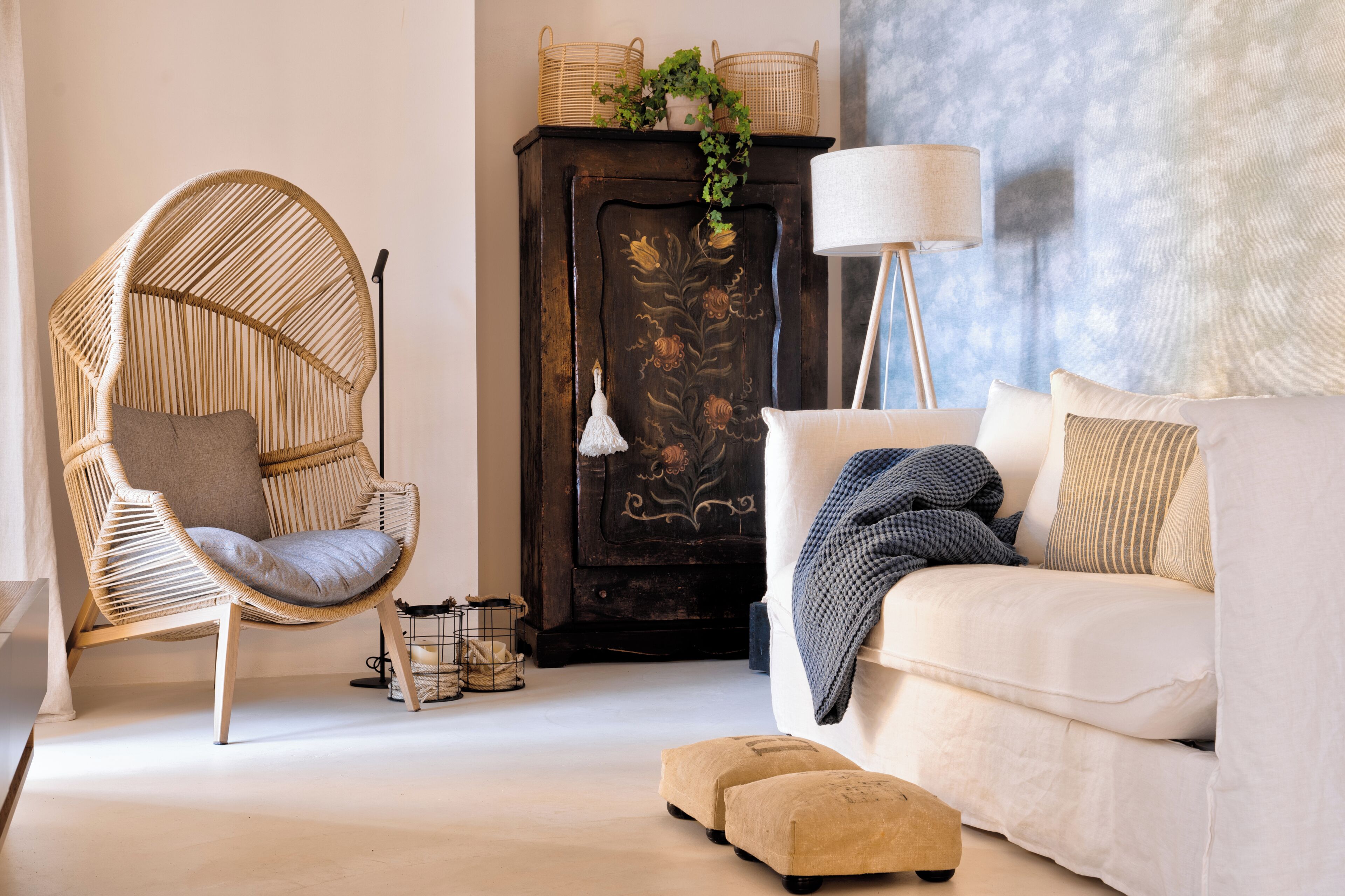 Ein Zimmer mit einem Rattan-Sessel, einer Couch, einem Schrank mit floralen Verzierungen und einer Stehlampe.