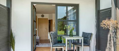 Een terras buiten met een tafel, twee stoelen, een bloempot en een deur die naar een binnenruimte leidt.