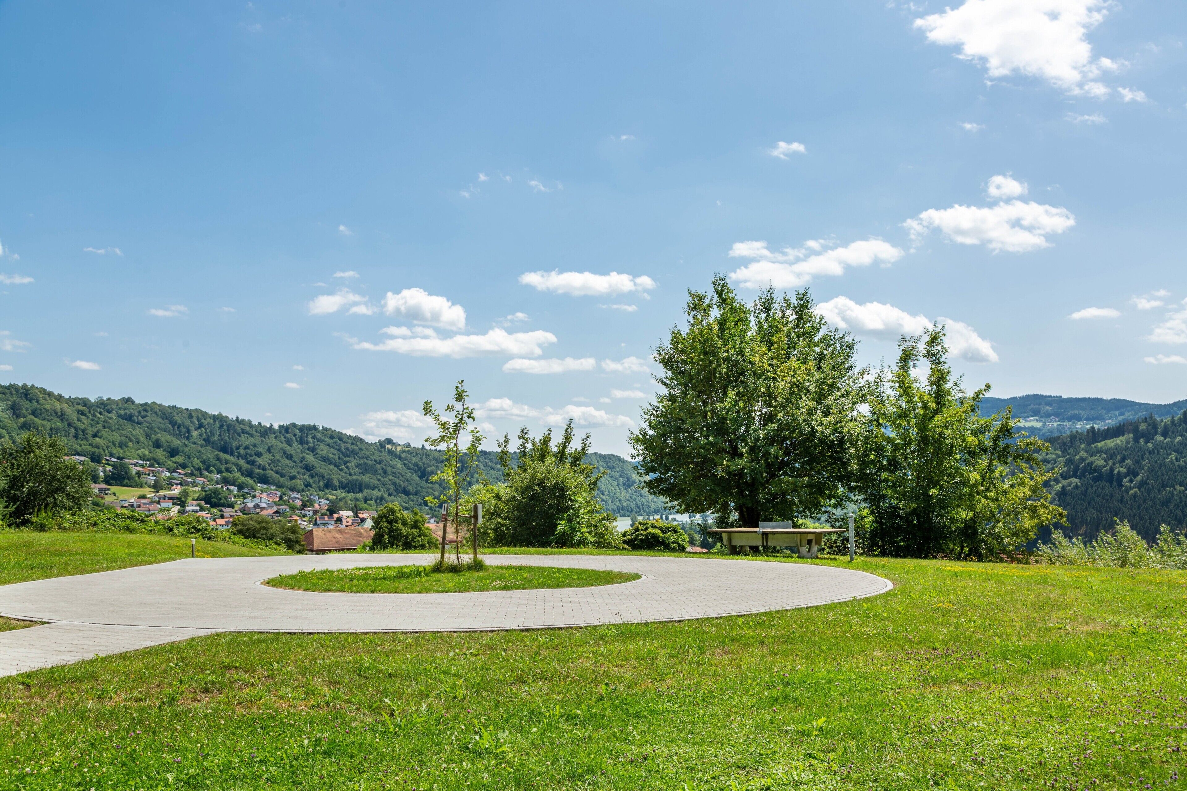 Ein gepflasterter Weg in einem Park mit einer Bank, umgeben von Gras und Bäumen, der zu einer Aussicht auf ein Dorf und Hügel in der Ferne führt.