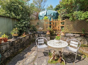 Eine Steinmauer, zwei Metallstühle, ein runder Metalltisch und Kübelpflanzen in einem Garten.