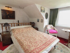 Ein Schlafzimmer mit einem Bett, einem Sessel, einem kleinen Tisch, einer Lampe, einem Gemälde und einem Fenster mit Vorhängen.