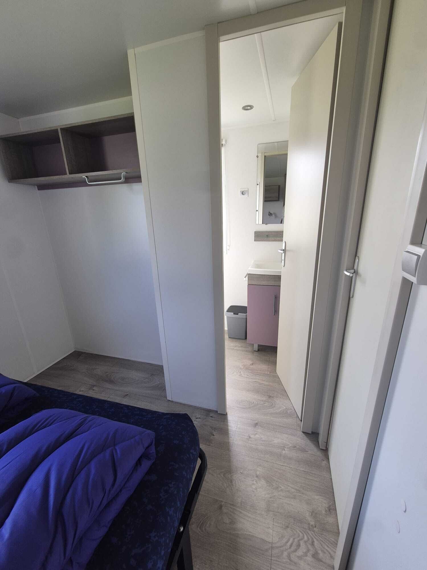 A bedroom with a bed, a wardrobe, a small bathroom with a mirror, and a trash bin.