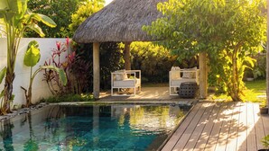 Un área de piscina con techo de paja, dos sillones blancos y una terraza de madera.