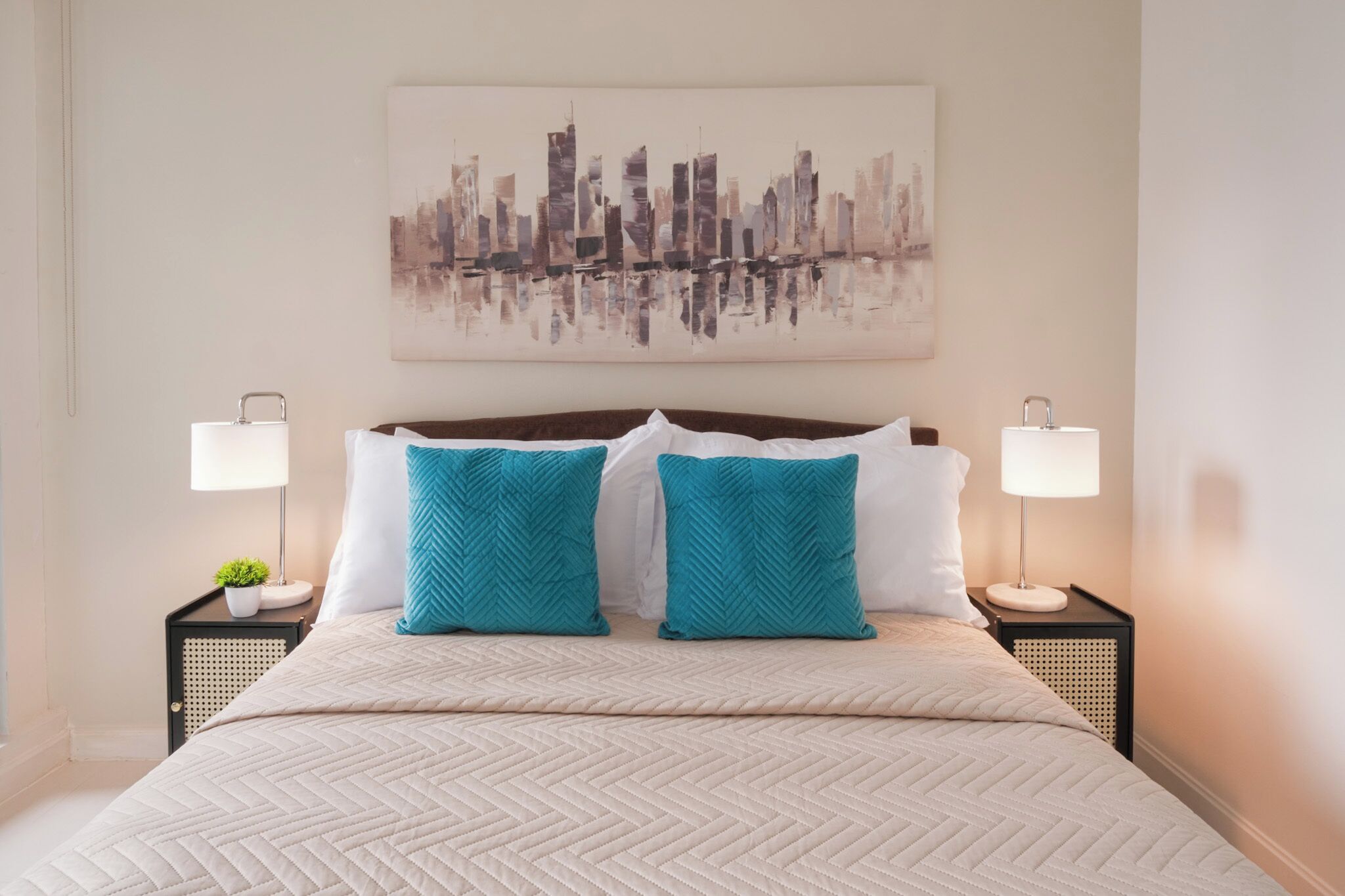 A neatly made bed with a patterned bedspread, two side tables with lamps, a small plant, and a cityscape painting on the wall.