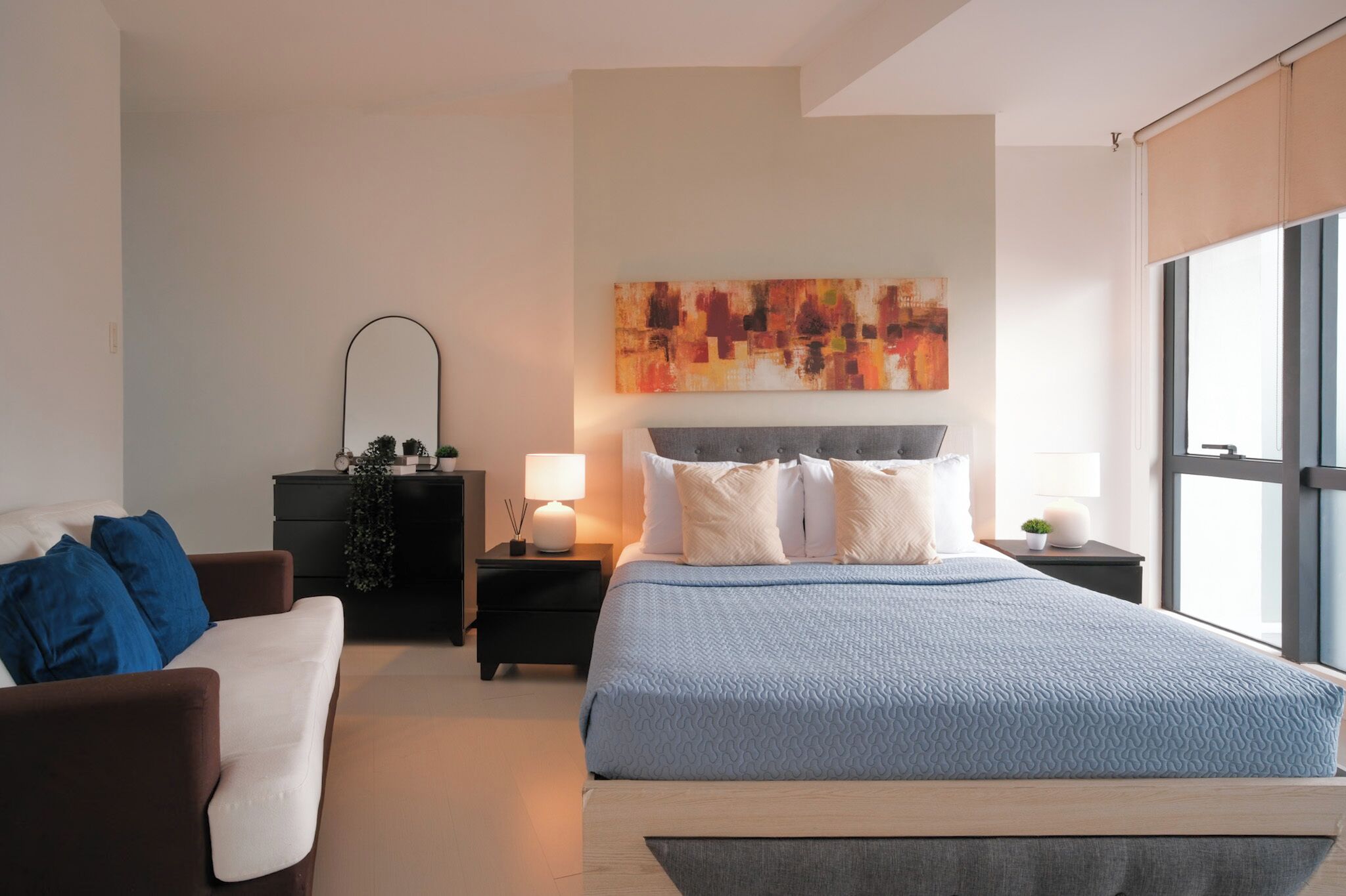 Une chambre à coucher moderne avec un grand lit, un canapé, une table de chevet avec une lampe, une commode et un grand tableau abstrait accroché au mur.