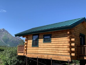 A wooden cabin with a green roof, two windows, and a deck. - Scenic Creekside Cabin with Panoramic Bay & Mountain Views in Seward, Alaska (Seward)