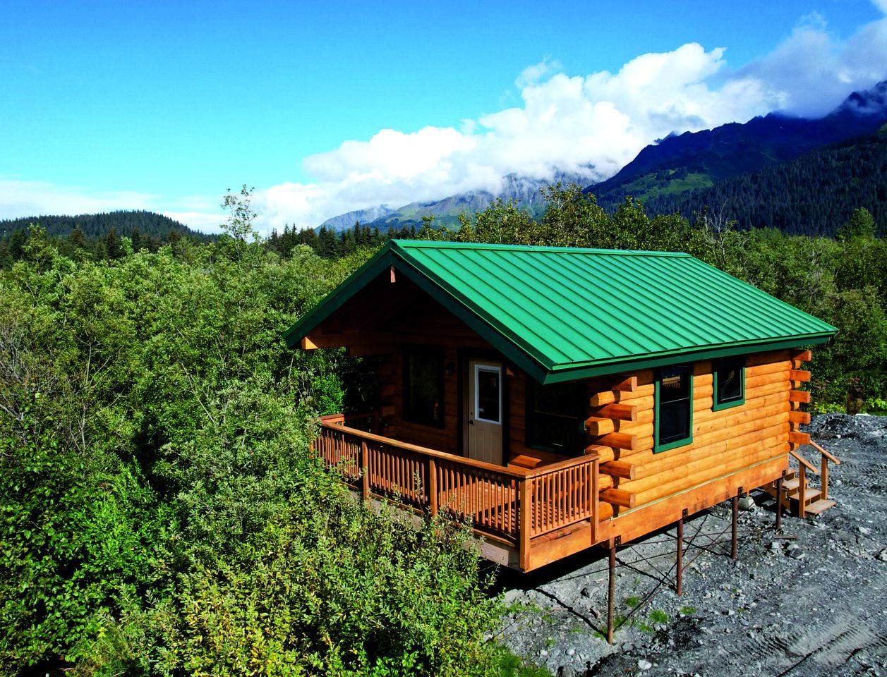 A wooden cabin with a green roof, surrounded by dense forest and mountains in the background.