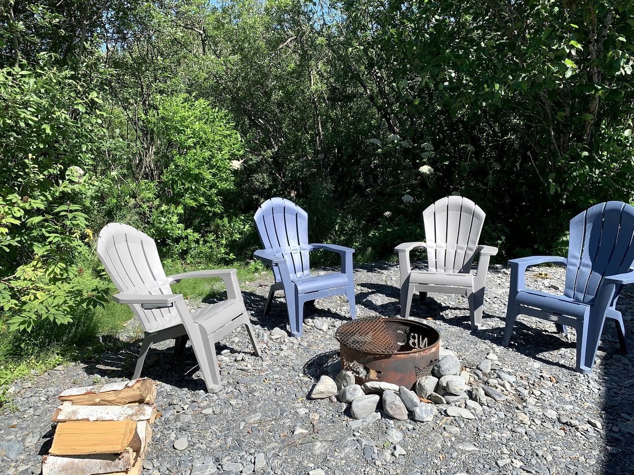 Vier Adirondack-stoelen staan rond een vuurkorf in een natuurlijke omgeving met bomen en rotsen.