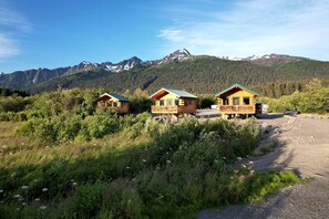 Trois chalets en bois dotés de balcons, entourés de verdure et avec des montagnes en toile de fond.