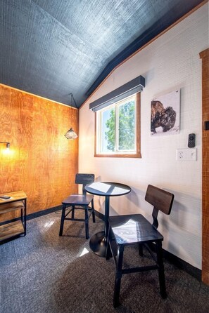 Ein Zimmer mit einer Holzwand, einem runden Tisch, zwei Stühlen, einer Bank, einem Fenster und einem Gemälde.
