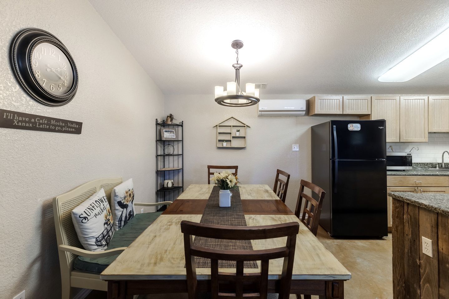 A dining area with a wooden table, chairs, a chandelier, a clock, and a refrigerator.