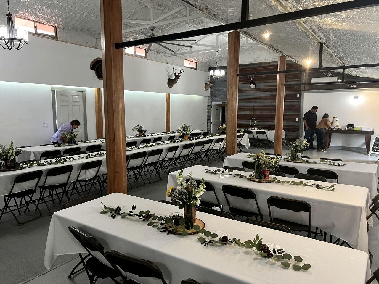 A dining area with tables set for a meal, a person arranging flowers, and another person standing by a counter.